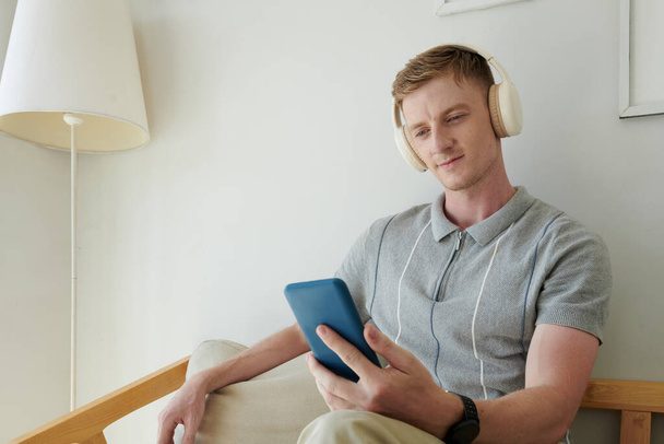 Joven sonriente en auriculares escuchando música de la lista de reproducción en el teléfono inteligente - Foto, Imagen