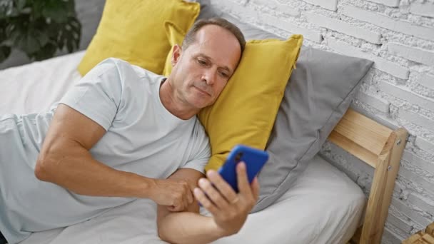 Starší, pohledný muž středního věku pohodlně uvolněný, ztracený ve svém smartphonu, intenzivně textuje, zatímco leží v posteli, přikrývky a pyžama na sobě, časně ráno ve svém teplém, útulném pokoji v ložnici - Záběry, video