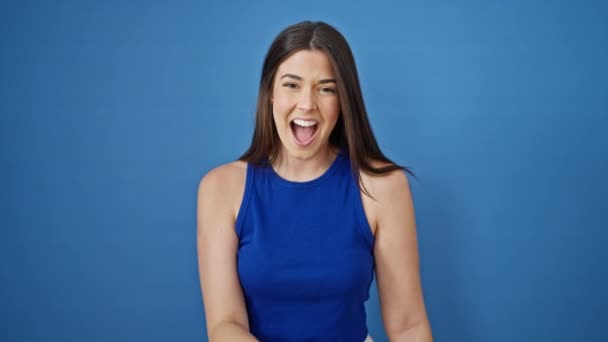 Fiatal gyönyörű spanyol nő áll a meglepetés kifejezés mutató kamera felett elszigetelt kék háttér - Felvétel, videó