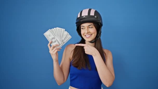 Молода красива іспаномовна жінка в шоломі мотоцикла вказує на долари, роблячи великий жест над ізольованим синім тлом - Кадри, відео