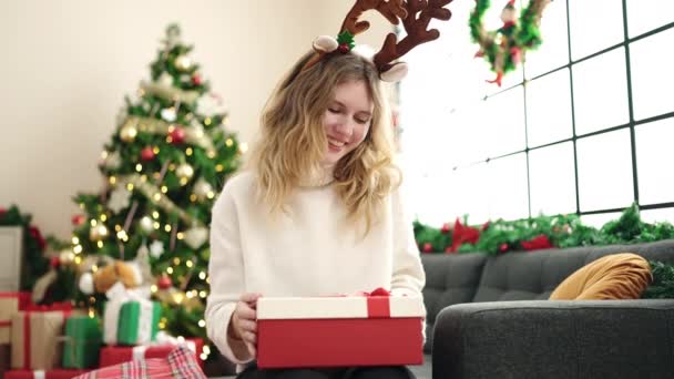 Jeune femme blonde déballer cadeau assis sur le canapé par arbre de Noël à la maison - Séquence, vidéo