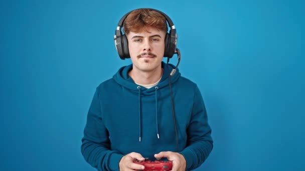 Junger hispanischer Mann spielt Videospiel und blickt verärgert über isolierten blauen Hintergrund - Filmmaterial, Video