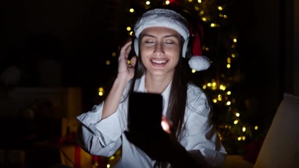 Νεαρή όμορφη ισπανόφωνη γυναίκα που χρησιμοποιεί το τηλέφωνο ακούγοντας μουσική να γιορτάζει τα Χριστούγεννα στο σπίτι - Πλάνα, βίντεο