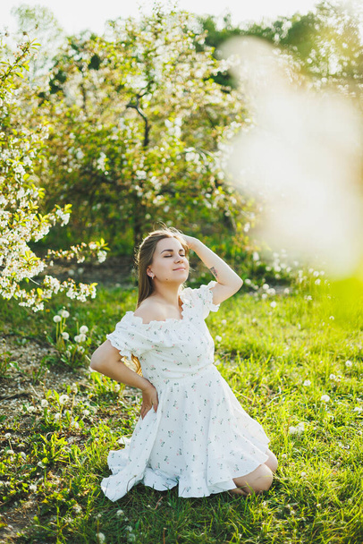 Eine glückliche schwangere Frau in einem sommerlichen weißen Musselinkleid sitzt auf dem Gras in einem blühenden Garten. Zukünftige Mutter, eine Frau, die ein Baby erwartet. Schwangerschaftszeit - Foto, Bild