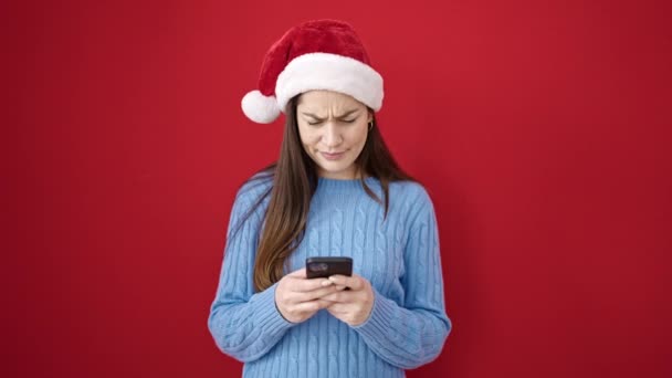 Junge kaukasische Frau trägt Weihnachtsmütze mit Smartphone vor isoliertem roten Hintergrund - Filmmaterial, Video
