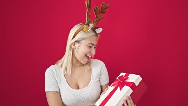 Młoda piękna Latynoska kobieta nosi renifery Boże Narodzenie kapelusz trzymając prezent nad odizolowanym czerwonym tle - Materiał filmowy, wideo