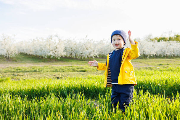 Egy mosolygó kisfiú sárga kabátban végigfut egy virágzó kerten. Boldog gyermekkort!. - Fotó, kép