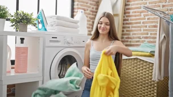 Jovem menina bonita sorrindo confiante jogando roupas no ar na lavanderia - Filmagem, Vídeo