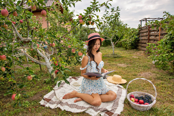 Όμορφη νεαρή μελαχρινή καυκάσιος γυναίκα φορώντας καπέλο κάθεται στον κήπο οπωρώνα και διαβάζοντας το βιβλίο. - Φωτογραφία, εικόνα