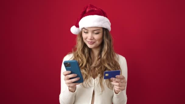 Joven mujer hispana hermosa usando sombrero de Navidad usando teléfono inteligente y tarjeta de crédito sobre fondo rojo aislado - Metraje, vídeo