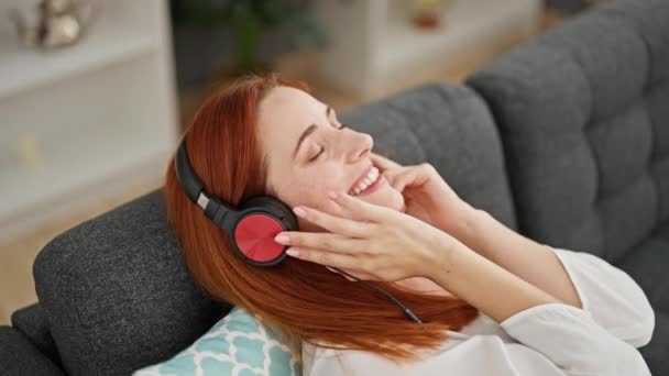 Mladá zrzka žena poslouchá hudbu sedí na pohovce doma - Záběry, video