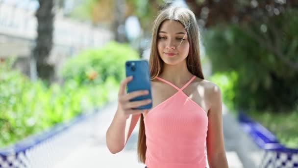 若い美しい女の子は自信を持って笑って公園のスマートフォンでセルフィーを作ります - 映像、動画