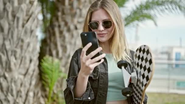Молодая блондинка держит скейт, используя смартфон в парке - Кадры, видео