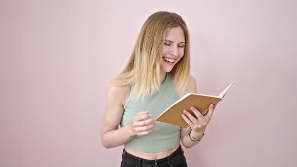 Νεαρή ξανθιά γυναίκα διαβάζει βιβλίο γελώντας πολύ πάνω σε απομονωμένο ροζ φόντο - Πλάνα, βίντεο