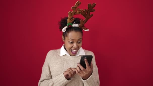 Afrika kökenli Amerikalı kadın, izole edilmiş kırmızı arka planda ren geyiği kulağı takarak akıllı telefon kullanıyor. - Video, Çekim