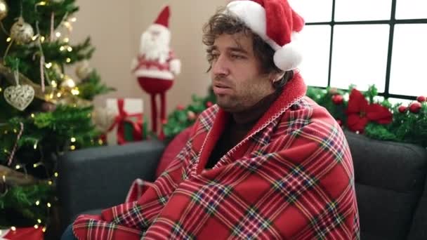 Young hispanic man feeling cold covered with blanket sitting on sofa by christmas tree at home - Footage, Video