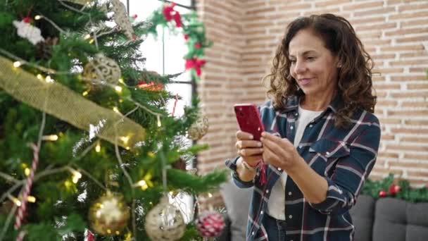 Mujer hispana de mediana edad grabando video en el árbol de navidad en casa - Imágenes, Vídeo