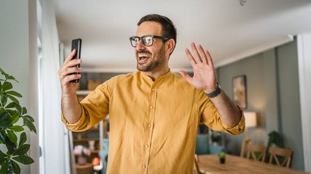 One man adult caucasian male with beard and eyeglasses stand at home happy smile use mobile phone smartphone to make a call talking video call copy space - Photo, Image