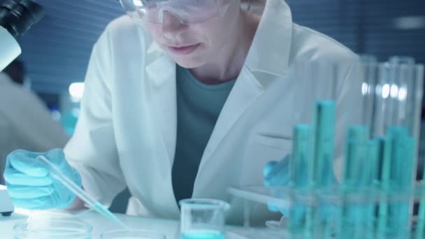 無菌手袋の女性化学者,顕微鏡下で化学物質を検査する保護眼鏡および白いコート - 映像、動画