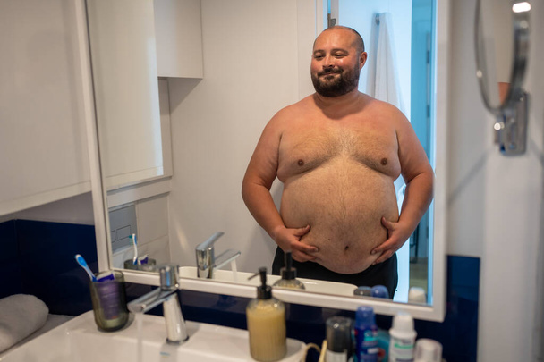 Χοντρός άντρας κοιτάζει τον καθρέφτη χαμογελώντας αγγίζοντας μεγάλη κοιλιά στο μπάνιο στο σπίτι. Ο γενειοφόρος δεν ικανοποιήθηκε με το σχήμα του σώματός του. παχυσαρκία, υπέρβαρο αρσενικό, επιπλέον βάρος, υγιή προβλήματα, έλεγχος βάρους. - Φωτογραφία, εικόνα