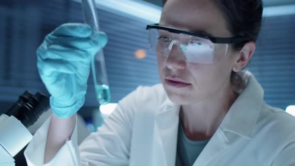 Химик-женщина в стерильных перчатках, защитных очках и белом халате изучает химическое вещество в пробирке во время работы в лаборатории - Кадры, видео