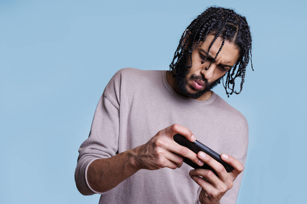 スマートフォンでオンラインゲームをプレイする集中アラブ男のゲーマー. 携帯電話を持っている若者と焦点を当てた表情の面白いモバイルアプリケーションソフトウェアを使用して - 写真・画像