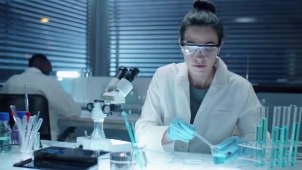 Tiro médio de mulher cientista em luvas estéreis, óculos de proteção e casaco branco examinando produtos químicos sob microscópio enquanto trabalhava tarde em laboratório - Filmagem, Vídeo