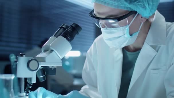 使い捨て手袋,帽子,顔面マスク,保護眼鏡を顕微鏡で見て研究室で働いている間にメモを作る女性科学者 - 映像、動画