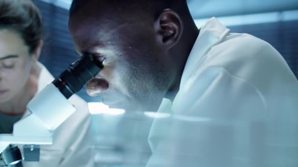 lähikuva Afrikkalainen amerikkalainen tiedemies katselee mikroskoopilla ja puhuu valkoihoinen naispuolinen avustaja, kun hän tekee muistiinpanoja aikana lab tutkimus - Materiaali, video