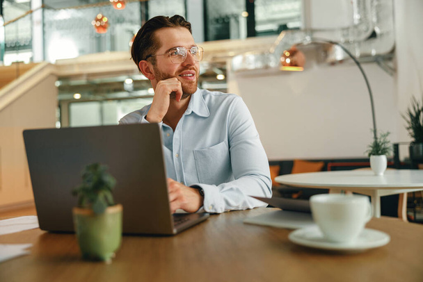 Uśmiechnięty menadżer pijący kawę podczas pracy na laptopie w nowoczesnym biurze. Wysokiej jakości zdjęcie - Zdjęcie, obraz