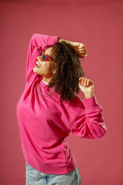 Smiling woman in sweater and heart-shaped glasses standing on pink studio background - Photo, Image