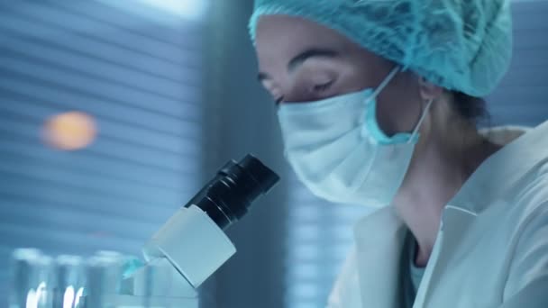 Portret van vrouwelijke wetenschapper in beschermend gezichtsmasker, steriele hoed en witte vacht met behulp van microscoop en dan poseren voor camera aan het werk in laboratorium - Video