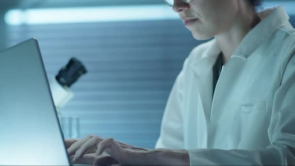 Tilt up shot of female scientist in lab coat and glasses working on laptop at desk in laboratory - Imágenes, Vídeo