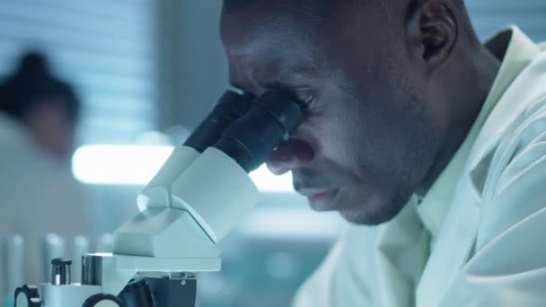Inyección de arco de primer plano del científico afroamericano en bata de laboratorio examinando la muestra a través del microscopio mientras investigaba en laboratorio - Imágenes, Vídeo