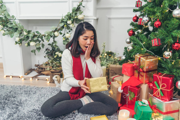 女性のサプライズはクリスマスイブにプレゼントギフトボックスを持って来ました. 笑顔のクリスマスツリーの下にクリスマスギフトボックスを持っているアジアの女性. 女性の手はプレゼントを包みます. メリークリスマスハッピーニューイヤーコンセプト - 写真・画像