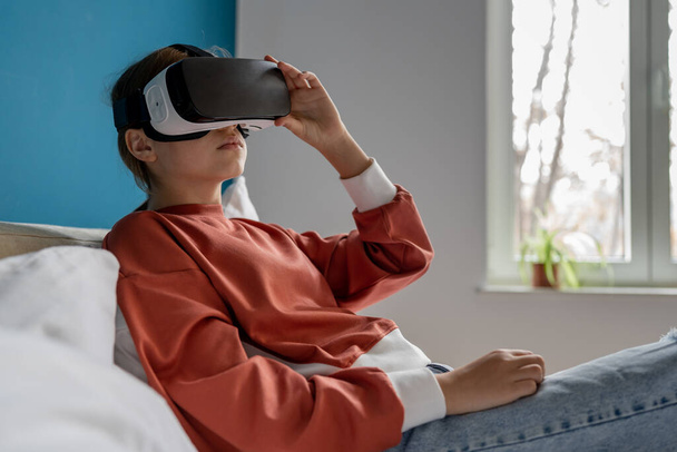 Relaxed teenage girl child in vr glasses watching 360-degree immersive educational video. Woman in VR goggles using metaverse headset lying on bed at home. Virtual reality, children and education - Photo, Image