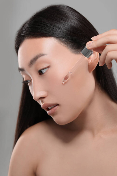 Beautiful young woman applying cosmetic serum onto her face on grey background - 写真・画像