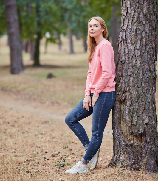 Niebieskooka blondynka w różowej bluzie spaceruje po lesie sosnowym. Portret radosnej młodej kobiety cieszącej się jesiennym parkiem. Relaks w przyrodzie. - Zdjęcie, obraz