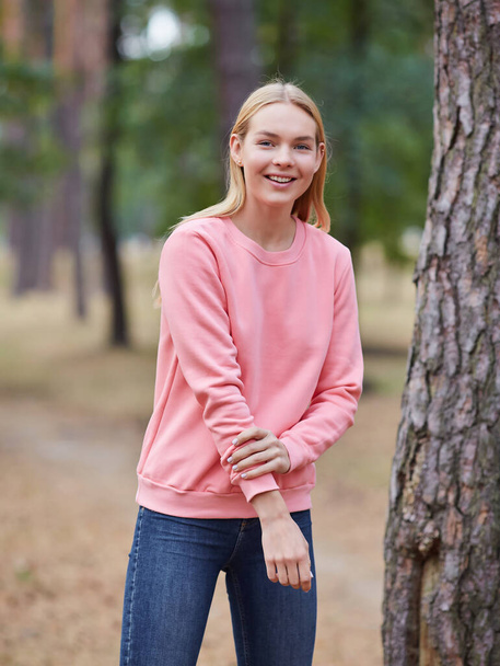 Приваблива блакитноока блондинка гуляє по міському парку. Дівчина носить рожевий светр, рожевий мішок і виглядає щасливою і посміхається. Портрет щасливої молодої жінки, яка насолоджується осіннім парком. Відпочинок в природі
. - Фото, зображення