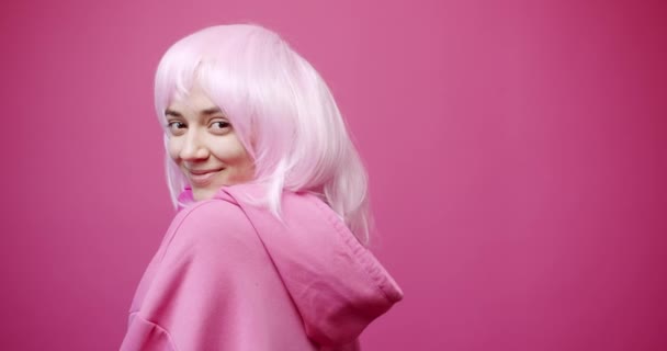 Fiatal nő flörtöl a kamerával. Statikus lövés boldog fiatal nő elegáns kapucnis vezeték nélküli fejhallgató és festett haj néz kamera mosollyal és kacsintott rózsaszín háttér - Felvétel, videó