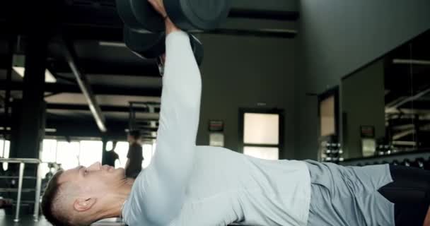Der junge Mann Bodybuilder trainiert im Fitnessstudio und hebt Hanteln, während er auf der Sportbank liegt. Athletischer Mann in Sportbekleidung beim Krafttraining für Brust- und Armmuskeln im Fitnessclub - Filmmaterial, Video
