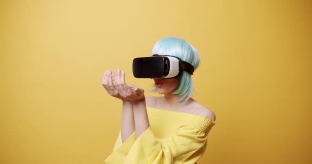 Mulher incrível explorando a realidade virtual. Tiro estático de fêmea jovem espantado com cabelo azul e capacete VR olhando ao redor e tocando objetos invisíveis contra fundo amarelo - Filmagem, Vídeo