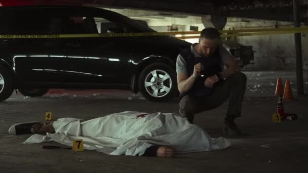 Full shot of Caucasian male police detective, in bulletproof vest and holding torch, investigating murder scene at dark underground parking lot and inspecting dead body covered with white sheet - Footage, Video