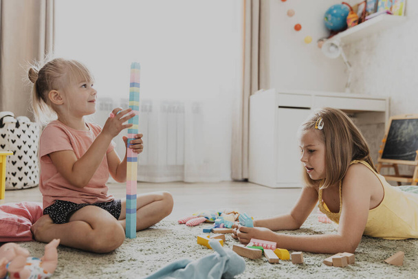 A gyerekek színes játékokkal játszanak. Gyerekek ülnek szőnyegen az otthoni hálószobában, óvodában, óvodában. Oktatási környezetbarát játék a gyermek számára. Imádnivaló kislányok építenek házat, tornyot együtt. - Fotó, kép