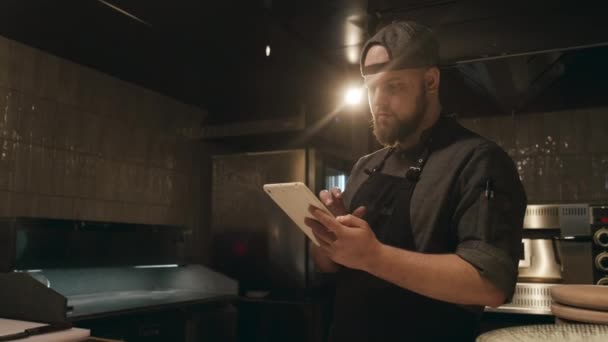 Średnie ujęcie młodego brodatego białego mężczyzny w mundurze szefa kuchni stojącego w słabo oświetlonej kuchni restauracyjnej, czekającego na zamówienia i korzystającego z tabletu - Materiał filmowy, wideo