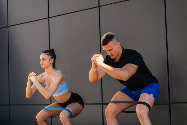 Handsome man and gorgeous woman, a couple, doing squats out in the city. They have wrapped resistance bands up around their legs - Foto, Imagen