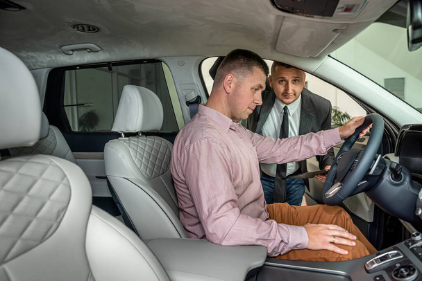 Jonge mannelijke cliënt zit in een nieuwe auto en praat met een competente manager in de showroom. concept van het verkoopvoertuig - Foto, afbeelding