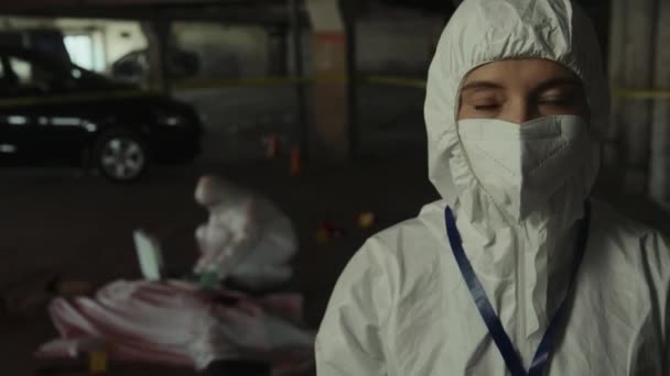 Medium close-up portrait shot of female forensic specialist in protective suit and face mask looking at camera with shock and stress, while colleague is collecting samples off corpse at crime scene - Footage, Video