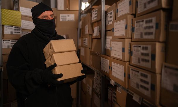 Un voleur dans un masque noir vole une boîte de marchandises dans un entrepôt la nuit. Problèmes liés au vol de marchandises et de colis postaux. Espace de copie - Photo, image
