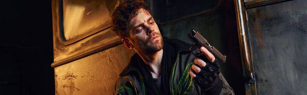 Mann mit verletztem Gesicht blickt auf Waffe in verlassener U-Bahn, postapokalyptischer Überlebender, Transparent - Foto, Bild